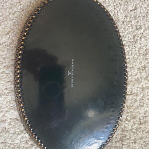 Puede incluir: Una bandeja ovalada de color negro con acabado brillante y un borde decorativo de estilo trenzado en oro met&aacute;lico. El centro de la bandeja presenta el texto MACKENZIE-CHILDS impreso en blanco con un peque&ntilde;o icono de logotipo debajo.