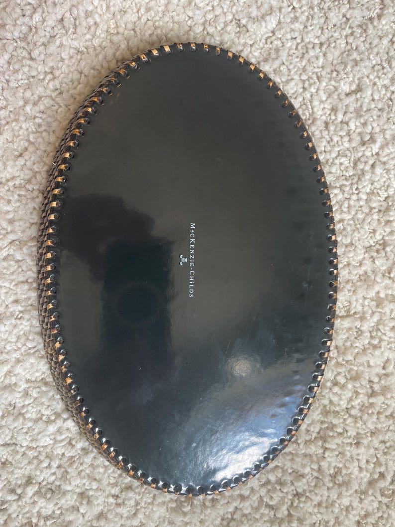 Puede incluir: Una bandeja ovalada de color negro con acabado brillante y un borde decorativo de estilo trenzado en oro met&aacute;lico. El centro de la bandeja presenta el texto MACKENZIE-CHILDS impreso en blanco con un peque&ntilde;o icono de logotipo debajo.