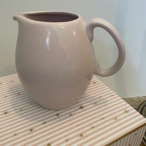 May include: A pale pink ceramic jug with a rounded body and a curved handle. The jug has a spout for easy pouring. It sits on a pink and white striped box with gold star accents. The jug is likely used for serving beverages.