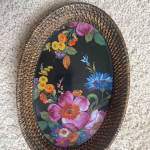 Puede incluir: Una bandeja decorativa de forma ovalada con un borde de rat&aacute;n trenzado marr&oacute;n con acabado en oro met&aacute;lico. El centro es una superficie negra lisa decorada con un estampado floral vibrante que muestra flores rosas, amarillas, naranjas y azules.