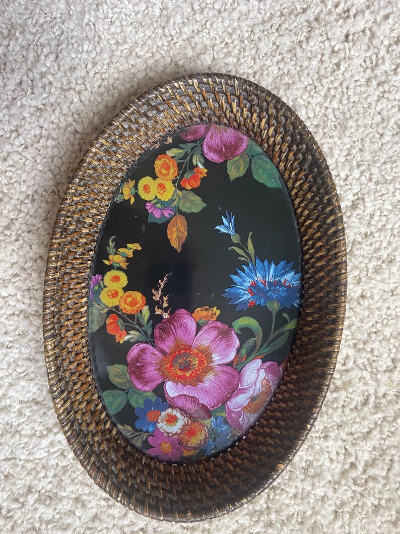 Puede incluir: Una bandeja decorativa de forma ovalada con un borde de rat&aacute;n trenzado marr&oacute;n con acabado en oro met&aacute;lico. El centro es una superficie negra lisa decorada con un estampado floral vibrante que muestra flores rosas, amarillas, naranjas y azules.
