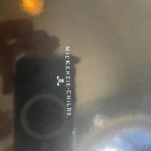 Puede incluir: Una superficie negra lisa y brillante con el texto blanco MACKENZIE-CHILDS impreso verticalmente en el centro, acompa&ntilde;ado de un peque&ntilde;o gr&aacute;fico blanco de una flor de cardo situado directamente encima del nombre de la marca.