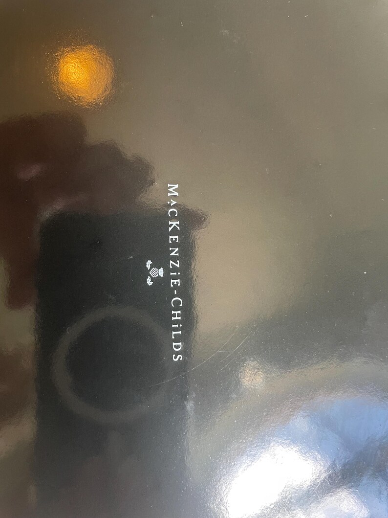 Puede incluir: Una superficie negra lisa y brillante con el texto blanco MACKENZIE-CHILDS impreso verticalmente en el centro, acompa&ntilde;ado de un peque&ntilde;o gr&aacute;fico blanco de una flor de cardo situado directamente encima del nombre de la marca.