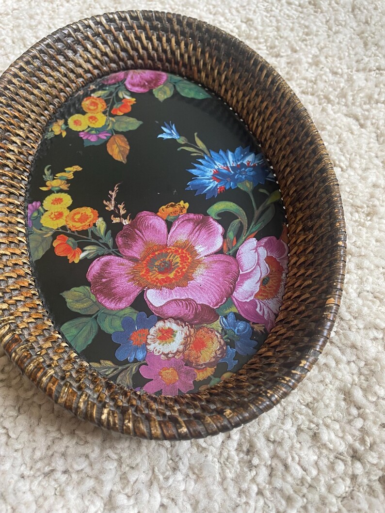 Puede incluir: Una bandeja de forma ovalada con un borde de rat&aacute;n tejido en color marr&oacute;n oscuro y una base plana decorada con un estampado floral vibrante sobre un fondo negro. El dise&ntilde;o floral incluye grandes flores rosas, azules, amarillas y naranjas.