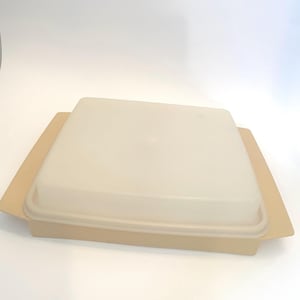May include: A vintage, square, beige plastic food storage container with a matching lid. The container has a slightly raised base and a flat, rectangular lid. The lid is a lighter shade of beige and fits snugly on top.