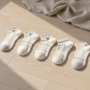 May include: Five pairs of white ankle socks with gray toes and various small embroidered designs. Each sock has a different design, including a bear, stripes, and other small patterns. The socks are arranged on a light-colored wooden surface.