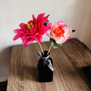 May include: A black glass vase holds two artificial flowers: a vibrant pink dahlia and a pink and orange rose. The vase is tied with a black and white ribbon. The arrangement sits on a wooden surface.