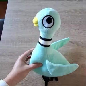 May include: A plush pigeon toy in shades of teal, with a yellow beak, black eyes, and a black and white collar. The stuffed animal has black feet and wings. The toy is being held by a person.