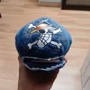 May include: A blue hat with a white and yellow graphic of a skull and crossbones with a straw hat. The hat has a white trim and is held in a person's hand.