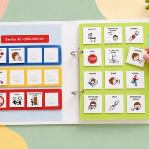 Könnte beinhalten: Ein weißer Ordner mit Kommunikationskarten, farbigen Streifen und quadratischen Kärtchen mit französischem Text und Illustrationen für tägliche Aktivitäten. Der Ordner enthält eine grüne Seite mit Registerkarten und eine Hand.