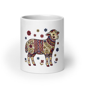 May include: White ceramic mug featuring a colorful illustration of a lamb with floral and geometric patterns. The lamb is decorated with red, blue, yellow, and brown designs. Surrounding the lamb are small flowers and stars in various colors.