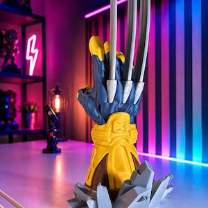 Può includere: Un pezzo da esposizione tridimensionale che presenta un guanto stilizzato blu e giallo con tre lunghe lame metalliche color argento che si estendono dalle nocche. Il guanto poggia su una base di frammenti rocciosi color argento.