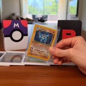 Puede incluir: Una carta coleccionable de Machamp en una funda protectora transparente, con la ilustraci&oacute;n de un personaje azul musculoso, 100 PV y el texto Movimento sismico 60. La carta se sostiene frente a una caja con dise&ntilde;o de Pok&eacute; Ball morado y blanco.