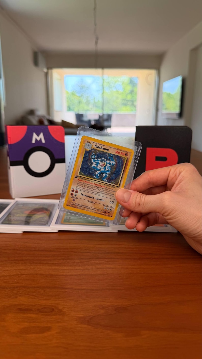 Puede incluir: Una carta coleccionable de Machamp en una funda protectora transparente, con la ilustraci&oacute;n de un personaje azul musculoso, 100 PV y el texto Movimento sismico 60. La carta se sostiene frente a una caja con dise&ntilde;o de Pok&eacute; Ball morado y blanco.
