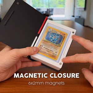 Puede incluir: Un estuche magn&eacute;tico de exhibici&oacute;n en blanco y negro que contiene una carta coleccionable Machamp. El estuche tiene una cubierta negra y un marco blanco con puntos de cierre magn&eacute;tico visibles. El texto dice MAGNETIC CLOSURE 6x2mm magnets.