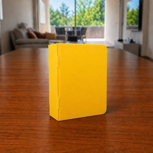 Puede incluir: Una caja rectangular de color amarillo s&oacute;lido con un panel lateral articulado. El objeto presenta un acabado mate liso y un mecanismo de bisagra vertical que conecta el cuerpo principal con el panel lateral, colocado sobre una superficie.