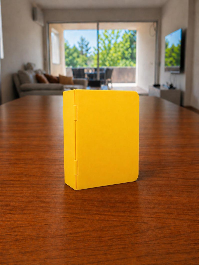 Puede incluir: Una caja rectangular de color amarillo s&oacute;lido con un panel lateral articulado. El objeto presenta un acabado mate liso y un mecanismo de bisagra vertical que conecta el cuerpo principal con el panel lateral, colocado sobre una superficie.