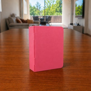 Puede incluir: Una carpeta rectangular de color rosa vibrante y s&oacute;lido con un acabado mate y suave. La carpeta presenta un lomo vertical en el lado izquierdo con dos bisagras visibles que permiten abrir la cubierta. El objeto se mantiene en posici&oacute;n vertical.