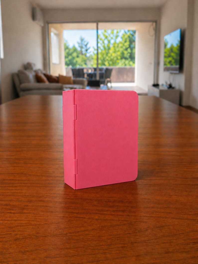 Puede incluir: Una carpeta rectangular de color rosa vibrante y s&oacute;lido con un acabado mate y suave. La carpeta presenta un lomo vertical en el lado izquierdo con dos bisagras visibles que permiten abrir la cubierta. El objeto se mantiene en posici&oacute;n vertical.