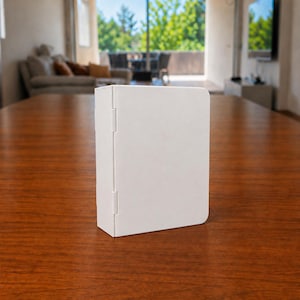 Puede incluir: Una carpeta rectangular de color blanco s&oacute;lido con un acabado mate suave y una bisagra vertical en el lado izquierdo. El objeto se mantiene en posici&oacute;n vertical sobre su borde, con un dise&ntilde;o limpio y esquinas redondeadas en el lado derecho.