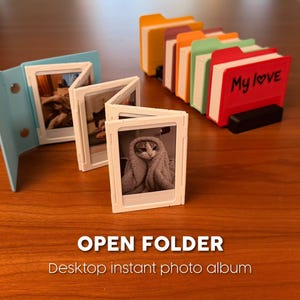 Puede incluir: Un álbum de fotos instantáneo de escritorio con una cubierta azul, que muestra varias fotos pequeñas de un gato. También se ve una fila de portafotos con forma de carpeta de colores. Se muestra el texto "OPEN FOLDER" y "Desktop instant photo album".