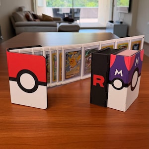 Puede incluir: Un archivador de cartas con tem&aacute;tica de Pok&eacute; Ball en blanco y rojo abierto para mostrar varias cartas coleccionables en fundas protectoras, junto a una caja de almacenamiento de Master Ball en blanco y negro con la letra R y la letra M.