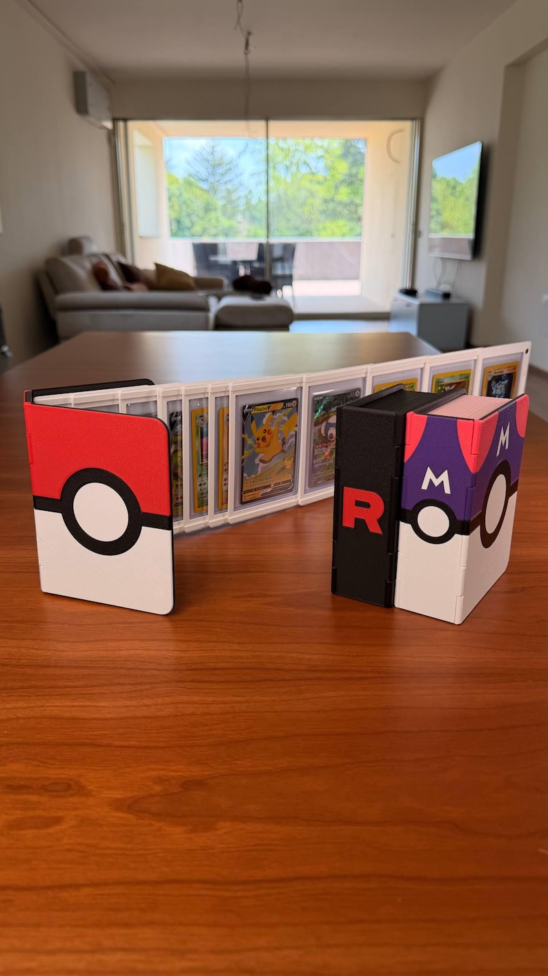 Puede incluir: Un archivador de cartas con tem&aacute;tica de Pok&eacute; Ball en blanco y rojo abierto para mostrar varias cartas coleccionables en fundas protectoras, junto a una caja de almacenamiento de Master Ball en blanco y negro con la letra R y la letra M.