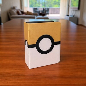 Puede incluir: Una caja de almacenamiento rectangular con una mitad superior dorada y una mitad inferior blanca, separadas por una banda negra horizontal con un emblema circular blanco y negro en el centro, que recuerda a una bola con esquinas redondeadas.