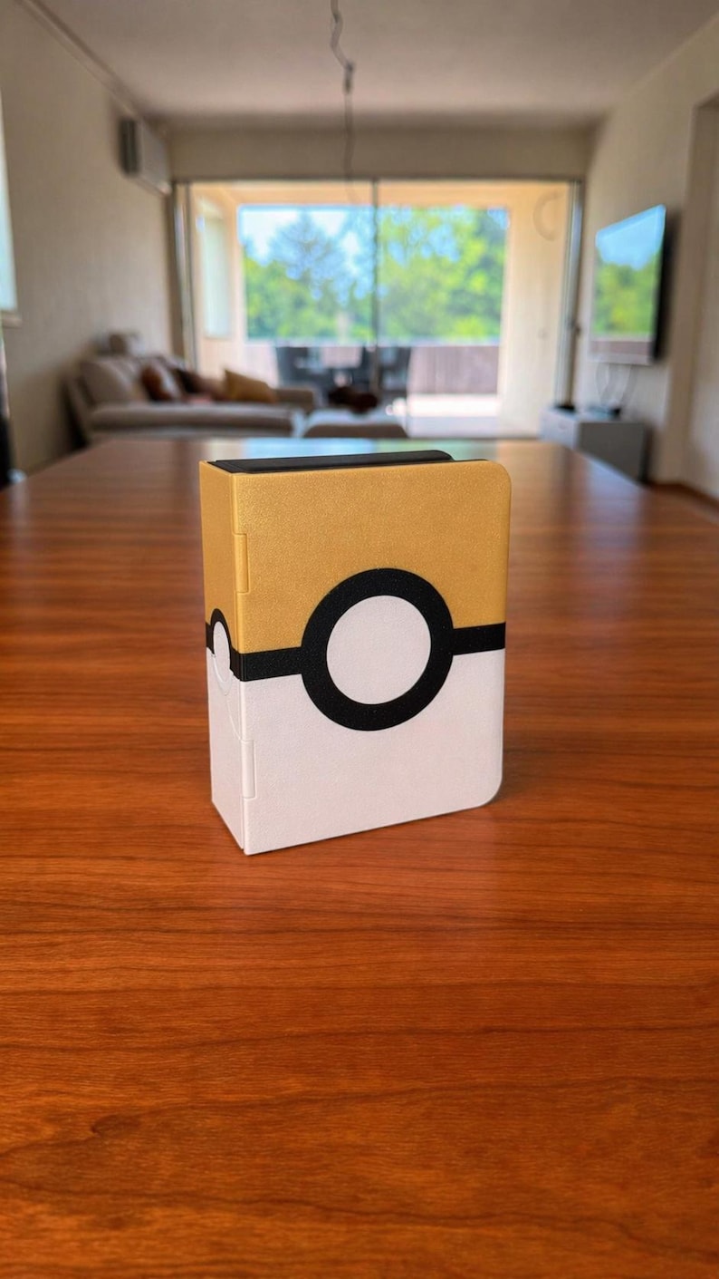 Puede incluir: Una caja de almacenamiento rectangular con una mitad superior dorada y una mitad inferior blanca, separadas por una banda negra horizontal con un emblema circular blanco y negro en el centro, que recuerda a una bola con esquinas redondeadas.