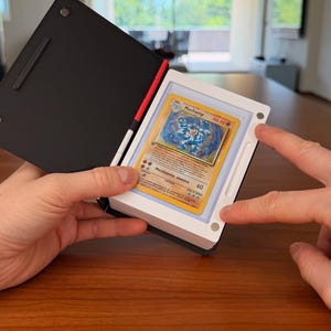 Puede incluir: Un estuche de exhibici&oacute;n magn&eacute;tico en blanco y negro que contiene una carta coleccionable de Machamp. El estuche tiene una cubierta negra y un marco blanco con cuatro peque&ntilde;os imanes circulares en las esquinas que aseguran la carta.