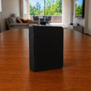 Puede incluir: Un estuche plegable rectangular de color negro mate con una superficie lisa y s&oacute;lida, y bisagras visibles en el lado izquierdo. El objeto se mantiene en posici&oacute;n vertical, con un dise&ntilde;o limpio, esquinas redondeadas y un acabado oscuro uniforme.