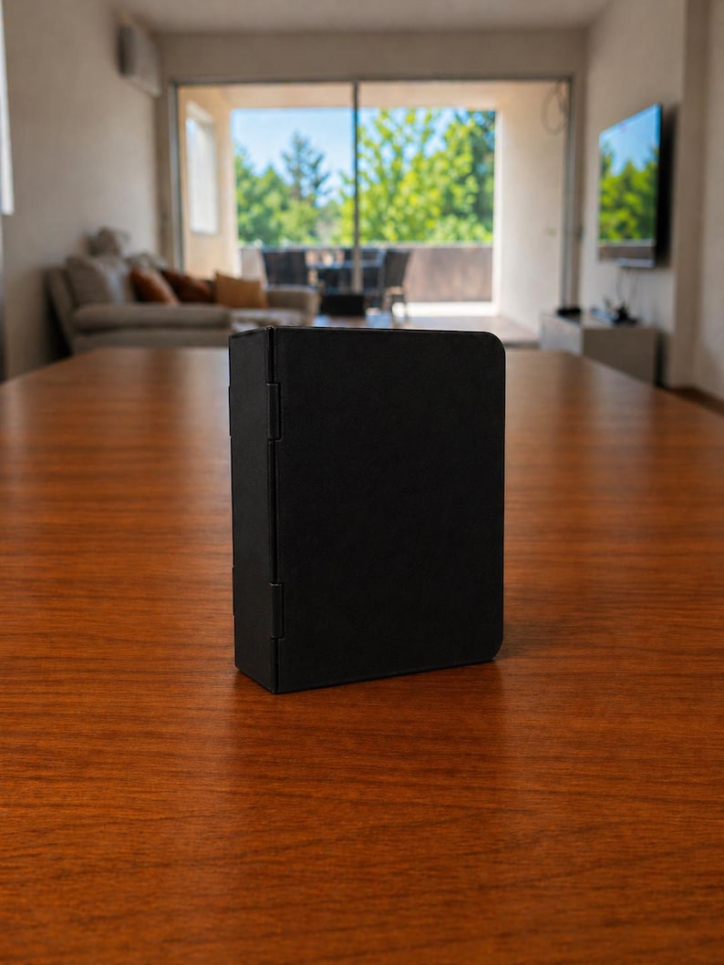 Puede incluir: Un estuche plegable rectangular de color negro mate con una superficie lisa y s&oacute;lida, y bisagras visibles en el lado izquierdo. El objeto se mantiene en posici&oacute;n vertical, con un dise&ntilde;o limpio, esquinas redondeadas y un acabado oscuro uniforme.