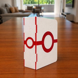 Puede incluir: Una caja rectangular blanca con superficie texturizada que presenta un gr&aacute;fico circular rojo brillante en la parte frontal y un patr&oacute;n de l&iacute;neas verticales rojas a lo largo del lomo. El objeto tiene un dise&ntilde;o geom&eacute;trico limpio y mate.