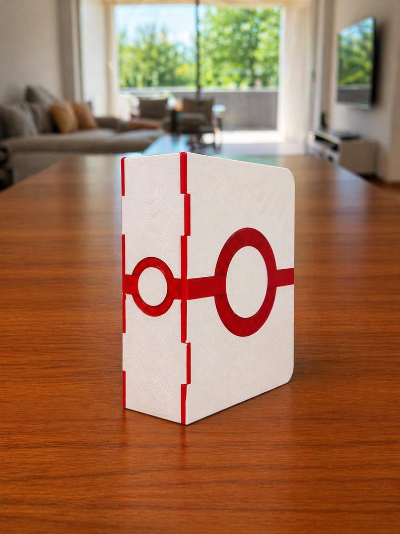 Puede incluir: Una caja rectangular blanca con superficie texturizada que presenta un gr&aacute;fico circular rojo brillante en la parte frontal y un patr&oacute;n de l&iacute;neas verticales rojas a lo largo del lomo. El objeto tiene un dise&ntilde;o geom&eacute;trico limpio y mate.