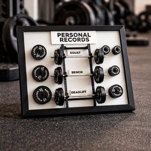 Peut inclure: Un tableau encadré avec les mots "PERSONAL RECORDS" en haut. Le tableau présente des haltères miniatures et des disques de poids pour les exercices de squat, de développé couché et de soulevé de terre. Les poids sont indiqués en kilogrammes.