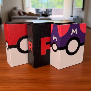 Puede incluir: Tres cajas de almacenamiento rectangulares colocadas en posici&oacute;n vertical. La caja de la izquierda es roja, blanca y negra con un emblema circular. La caja central es negra con una letra R roja. La derecha es morada, roja, blanca y negra con dos M.
