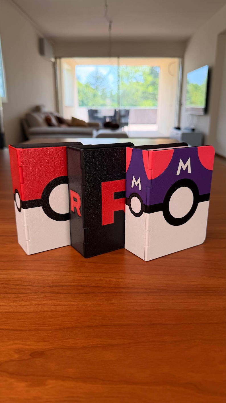 Puede incluir: Tres cajas de almacenamiento rectangulares colocadas en posici&oacute;n vertical. La caja de la izquierda es roja, blanca y negra con un emblema circular. La caja central es negra con una letra R roja. La derecha es morada, roja, blanca y negra con dos M.