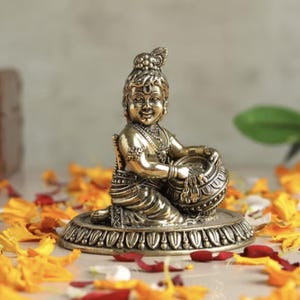 Puede incluir: Una figura de latón de una deidad sentada, sosteniendo una olla. La figura está detallada con ropa y joyas ornamentadas. La figura se asienta sobre una base decorativa, rodeada de pétalos de flores naranjas. El tema general es religioso.