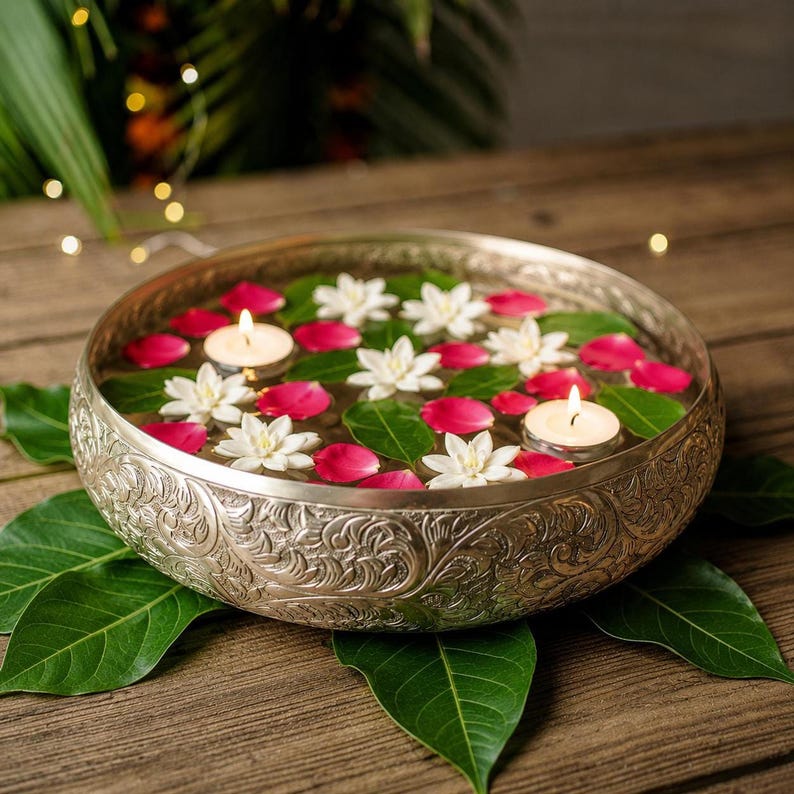 An ornate gold metal bowl filled with water, floating white flowers, red petals, and two lit tealight candles, resting on a wooden surface with green leaves.