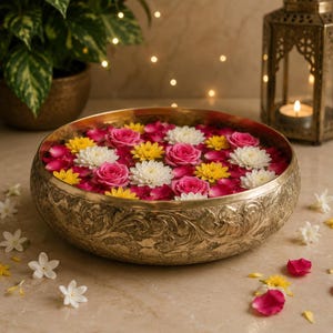 A decorative gold metal bowl filled with water, floating pink, white, and yellow flowers, and scattered petals, set on a surface with a potted plant and lantern in the background.
