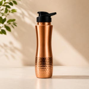 Op de afbeelding: Een koperkleurige metalen waterfles met een glad bovenste gedeelte en een gehamerde textuur op de onderste helft. De fles is voorzien van een zwarte kunststof dop met een klapdeksel en een handvat om de fles gemakkelijk te dragen.