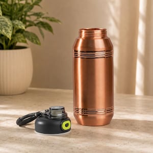 Op de afbeelding: Een koperkleurige metalen waterfles met een cilindrisch lichaam en horizontale ribbeldetails bij de boven- en onderkant. De fles wordt vergezeld door een losse zwarte plastic schroefdop met een limoengroene knop.