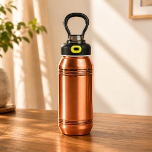 Op de afbeelding: Een koperkleurige metalen waterfles met een cilindrisch lichaam voorzien van horizontale ribbels. De fles heeft een zwarte schroefdop met een limoengroene accentknop en een grote, ge&iuml;ntegreerde zwarte draagbeugel aan de bovenzijde.