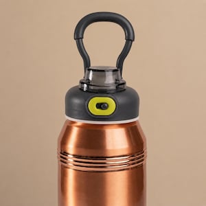 Op de afbeelding: Een koperkleurige metalen drinkfles met een donkergrijze schroefdop en een ge&iuml;ntegreerde draagbeugel. De dop is voorzien van een limoengroen schuifmechanisme en een doorzichtige tuitafdekking, met twee horizontale ribbels op de hals.