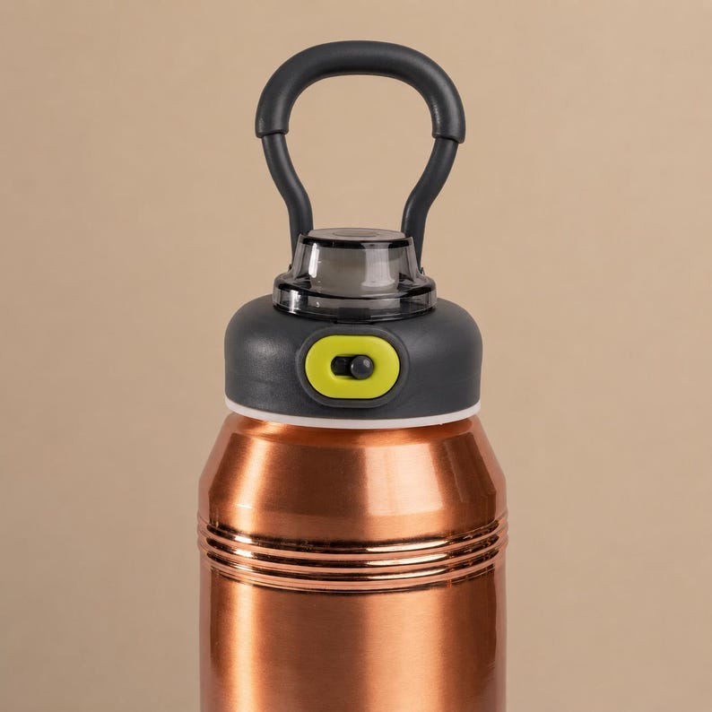 Op de afbeelding: Een koperkleurige metalen drinkfles met een donkergrijze schroefdop en een ge&iuml;ntegreerde draagbeugel. De dop is voorzien van een limoengroen schuifmechanisme en een doorzichtige tuitafdekking, met twee horizontale ribbels op de hals.