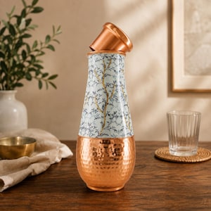 May include: A tall, tapered water bottle featuring a white upper section decorated with a delicate blue and gold branch pattern. The lower portion is crafted from hammered copper with a metallic finish, and it includes a matching copper cap.