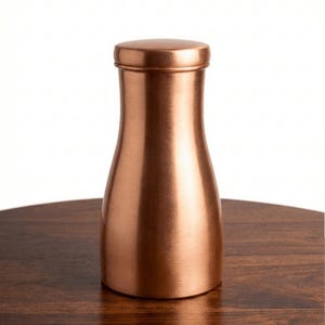 May include: A copper-colored, cylindrical container with a rounded lid. The container has a narrow neck and a wider base, sitting on a dark wooden surface. The metallic finish reflects light, highlighting its smooth surface.