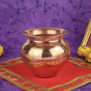 May include: A copper-colored pot with a wide rim, reflecting light, sits on a red cloth with a gold border. The background is a textured purple fabric. The pot is likely used for religious or ceremonial purposes.