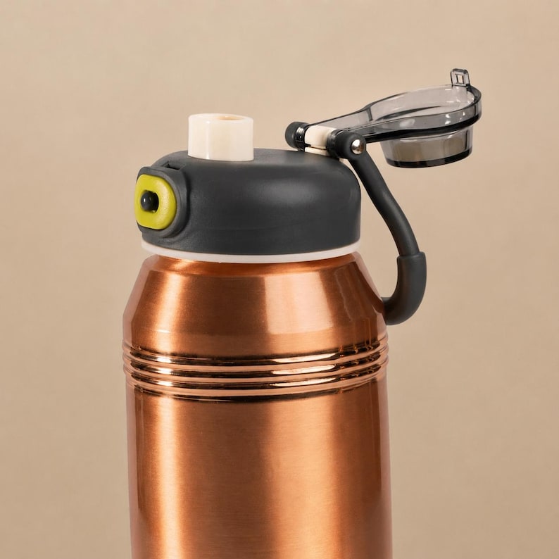 Op de afbeelding: Een koperkleurige metalen waterfles met een donkergrijze dop. De dop is voorzien van een wit mondstuk, een limoengroene knop en een scharnierende, doorschijnende donkergrijze klep die vastzit aan een flexibele donkergrijze band.
