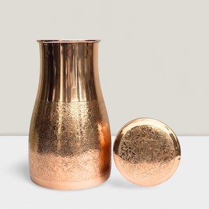 May include: A copper-colored water pitcher with an ornate, engraved floral pattern. The pitcher has a wide base that tapers to a narrower neck. The lid, also copper-colored, is placed next to the pitcher and features the same engraved design.